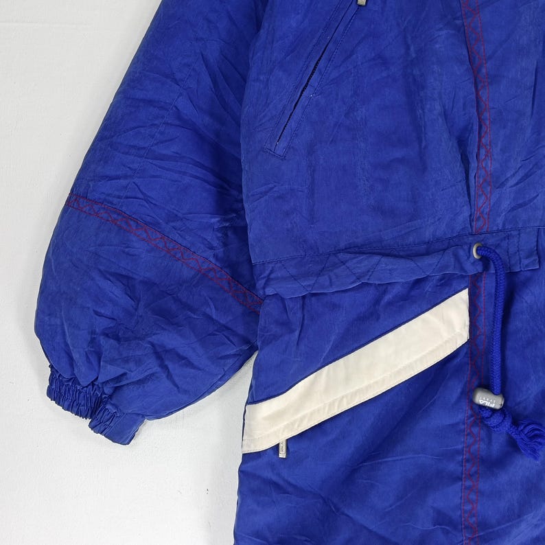 May include: Close-up of a blue and white vintage ski jacket. The jacket features a drawstring waist, a zippered pocket, and red stitching accents. The jacket has a white diagonal stripe and a blue drawstring.