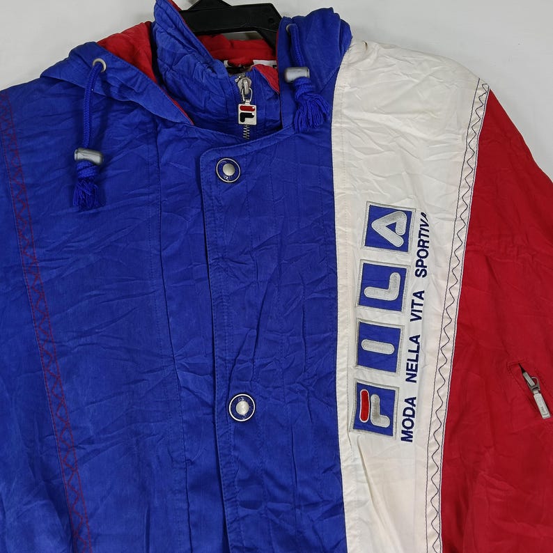 May include: A blue, red, and white FILA jacket with a hood. The jacket has a zipper and buttons. The FILA logo is displayed on the front. The jacket is made of a lightweight material.