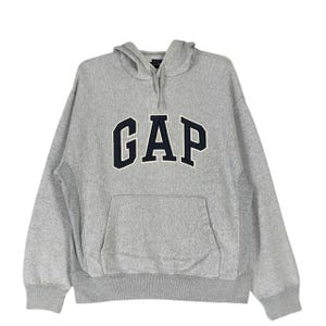 Può includere: Felpa con cappuccio grigio chiaro con un grande logo "GAP" blu navy sul petto. Ha una tasca a marsupio e un cappuccio con coulisse. Il tessuto sembra morbido e confortevole.