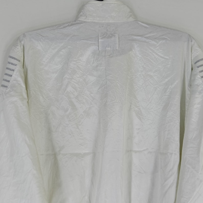 May include: A white, full-zip jacket with a high collar. The jacket has a subtle, textured pattern across the fabric. The shoulders feature three parallel gray stripes. The jacket is made of a lightweight material.