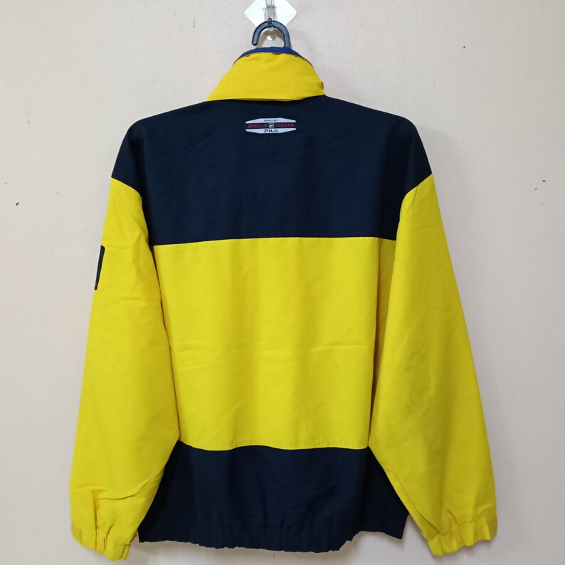 Puede incluir: Chaqueta Fila vintage en azul marino y amarillo. La chaqueta presenta un dise&ntilde;o de bloques de color con una secci&oacute;n superior azul marino y una secci&oacute;n media amarilla. El logotipo de Fila es visible en la parte superior trasera. La chaqueta tiene cremallera completa y cuello alto.