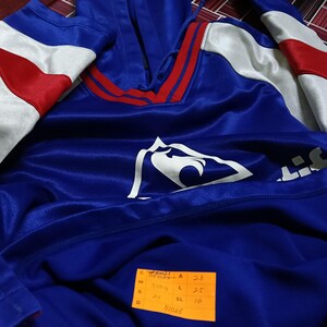 K&ouml;nnte beinhalten: Blauer Kapuzenpullover mit einem wei&szlig;en Adlerlogo und roten und wei&szlig;en Streifen an den &Auml;rmeln. Der Pullover hat ein Gr&ouml;&szlig;enetikett mit folgenden Ma&szlig;en: C 22, W 52, S 31, D 16, A 23, L 25, SL 16. Das Etikett hat auch die Nummer 11023.