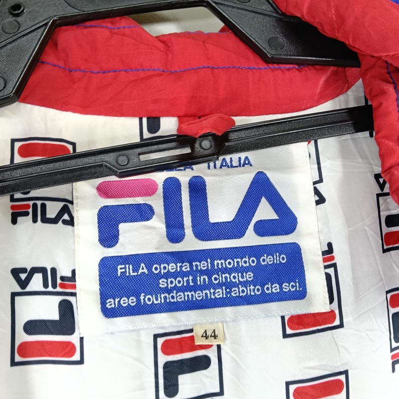May include: Close-up of a white and red Fila jacket with the Fila logo prominently displayed. The jacket has a red collar and a white interior with a repeating Fila logo pattern. The tag reads "FILA opera nel mondo dello sport in cinque aree foundamental: abito da sci."
