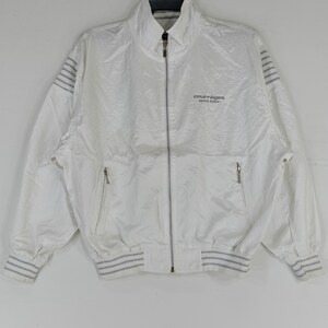 May include: White zip-up jacket with a high collar and silver zipper. The jacket features two front pockets and grey striped accents on the cuffs and shoulders. The Courr&egrave;ges logo is printed on the chest.