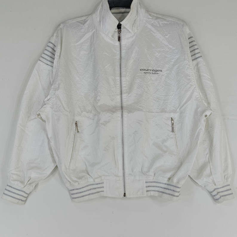 May include: White zip-up jacket with a high collar and silver zipper. The jacket features two front pockets and grey striped accents on the cuffs and shoulders. The Courr&egrave;ges logo is printed on the chest.