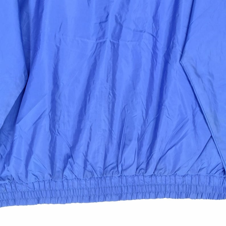May include: Close-up of a solid periwinkle blue jacket. The jacket has a gathered elastic waistband and a smooth, slightly shiny texture. The fabric is creased, suggesting it is made of a lightweight material.