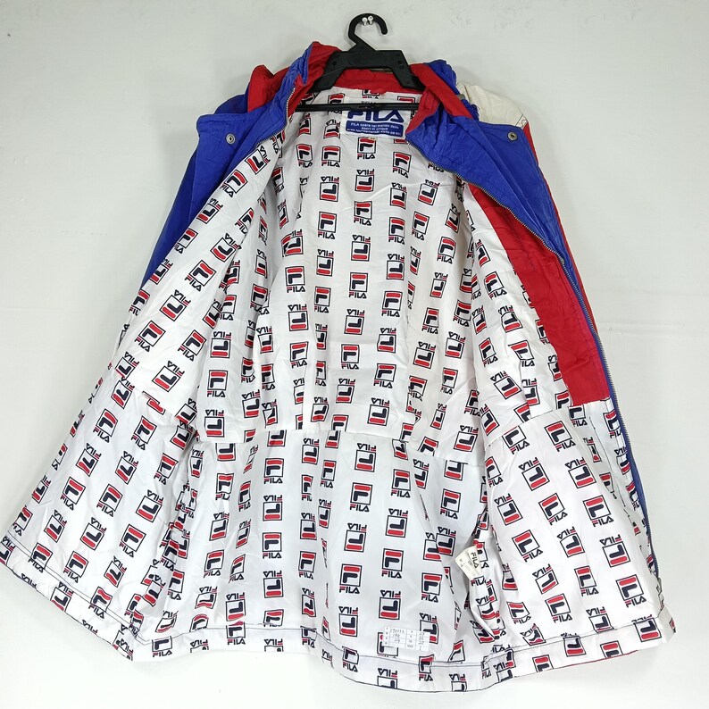 May include: A vintage Fila jacket in blue, red, and white. The jacket has a hood and is open, revealing a white lining with a repeating Fila logo pattern. The jacket is hanging on a black hanger.