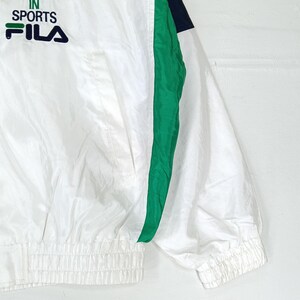 May include: White athletic jacket with green and navy blue accents. The jacket features the text "IN SPORTS FILA" in green and navy blue. The jacket has a ribbed hem and cuffs, and a front pocket.