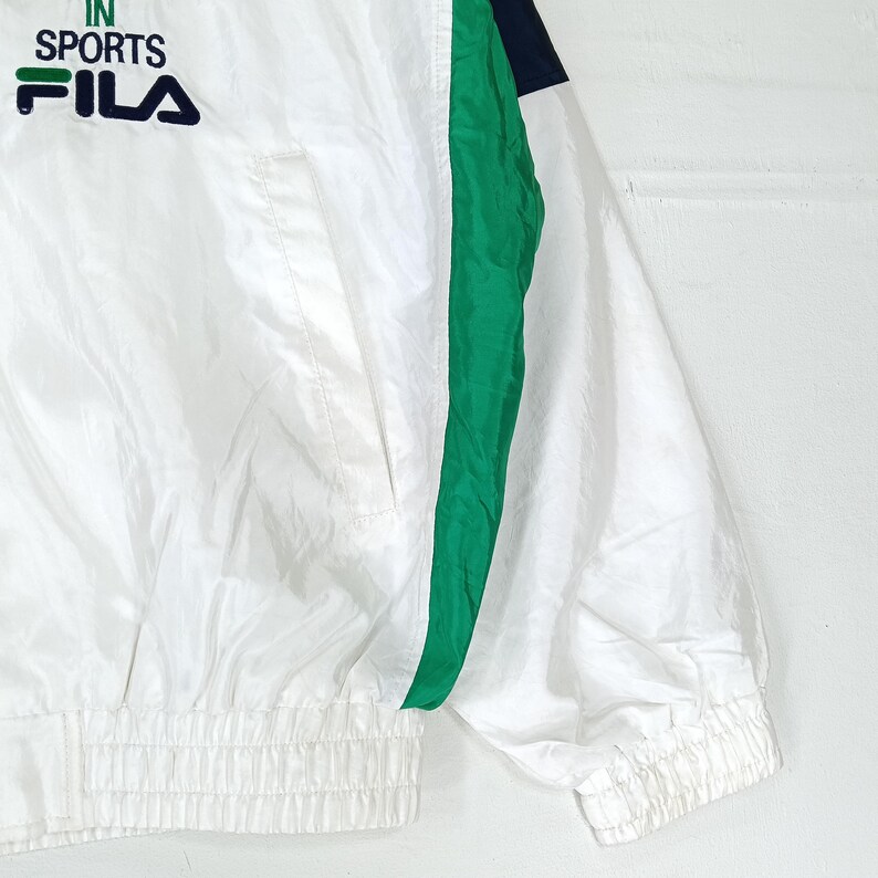 May include: White athletic jacket with green and navy blue accents. The jacket features the text "IN SPORTS FILA" in green and navy blue. The jacket has a ribbed hem and cuffs, and a front pocket.