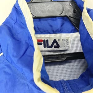 May include: A blue Fila jacket with white and cream accents. The jacket has a zipper closure and a tag with the Fila logo and size information. The jacket is hanging on a black hanger.