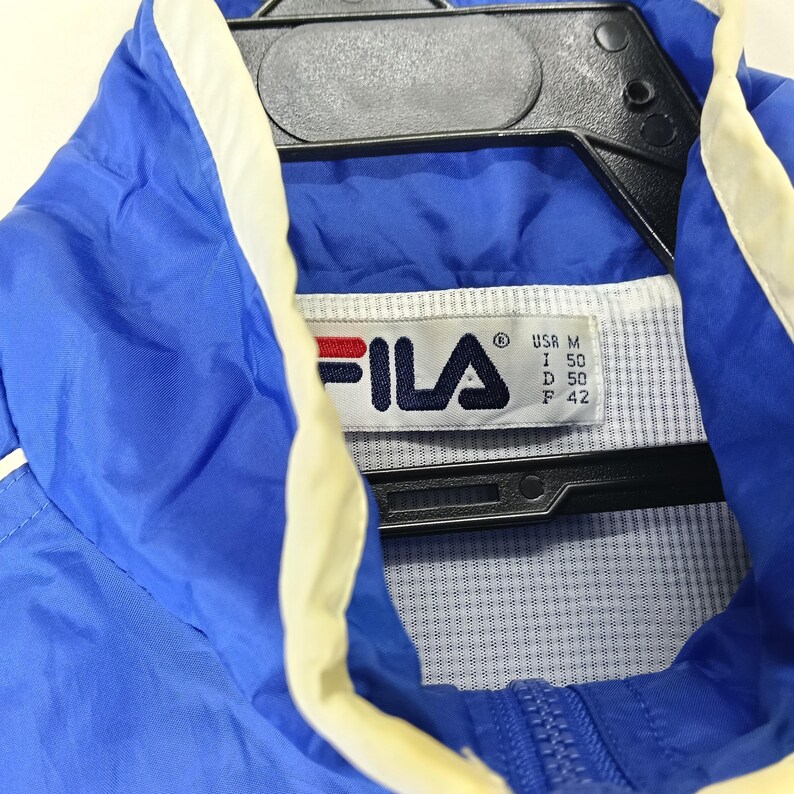 May include: A blue Fila jacket with white and cream accents. The jacket has a zipper closure and a tag with the Fila logo and size information. The jacket is hanging on a black hanger.