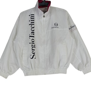 Op de afbeelding: Witte windbreaker-jas met ritssluiting en een donkerblauw logo met de tekst "Sergio Tacchini" op de voorkant. De jas heeft een roze voering en een donkerblauwe elastische tailleband.