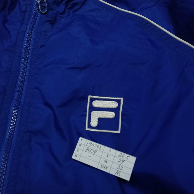 May include: A blue nylon jacket with a white trim and a full zipper. The jacket features the Fila logo on the chest. A tag with the numbers "190222" and "4009" is visible.