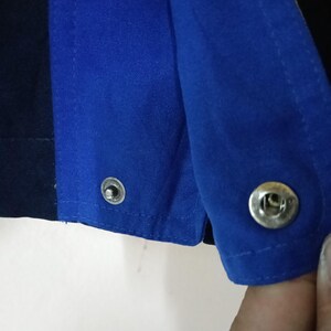 Puede incluir: Primer plano de una prenda azul y azul marino con cierres de broche plateados. La tela parece ser un material resistente, posiblemente de ropa de trabajo. La imagen destaca los detalles de las costuras y los broches met&aacute;licos.