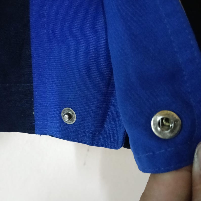 Puede incluir: Primer plano de una prenda azul y azul marino con cierres de broche plateados. La tela parece ser un material resistente, posiblemente de ropa de trabajo. La imagen destaca los detalles de las costuras y los broches met&aacute;licos.