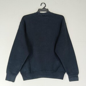K&ouml;nnte beinhalten: Ein marineblaues Sweatshirt h&auml;ngt an einem schwarzen Kleiderb&uuml;gel. Das Sweatshirt hat einen Rundhalsausschnitt und lange &Auml;rmel. Der Stoff scheint ein weiches, bequemes Material zu sein. Das Kleidungsst&uuml;ck wird vor einem schlichten wei&szlig;en Hintergrund pr&auml;sentiert.