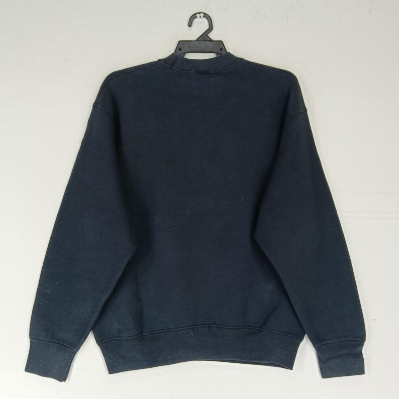 K&ouml;nnte beinhalten: Ein marineblaues Sweatshirt h&auml;ngt an einem schwarzen Kleiderb&uuml;gel. Das Sweatshirt hat einen Rundhalsausschnitt und lange &Auml;rmel. Der Stoff scheint ein weiches, bequemes Material zu sein. Das Kleidungsst&uuml;ck wird vor einem schlichten wei&szlig;en Hintergrund pr&auml;sentiert.