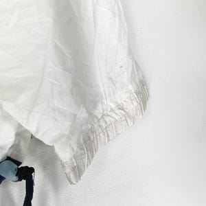 May include: Close-up of a white garment with a textured, slightly wrinkled appearance. The edge of the garment features a light gray trim. A dark blue drawstring and a blue and white plastic piece are visible at the bottom.