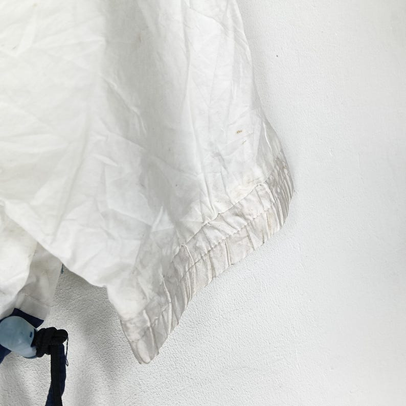 May include: Close-up of a white garment with a textured, slightly wrinkled appearance. The edge of the garment features a light gray trim. A dark blue drawstring and a blue and white plastic piece are visible at the bottom.