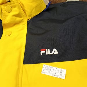 Puede incluir: Chaqueta amarilla y azul marino con el logo de FILA en blanco y rojo. La chaqueta tiene un cuello abotonado y una etiqueta con informaci&oacute;n de tallas. La chaqueta est&aacute; hecha de un material duradero.