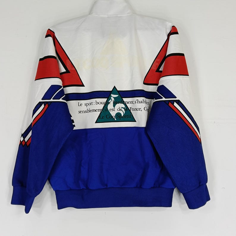 May include: A white, blue, and red jacket with a zipper closure. The jacket has a geometric pattern and the text "Le sport: bouge d'un mouvement, s'habille sensiblement, est de la France, C'est Le Coq Sportif" on the back.