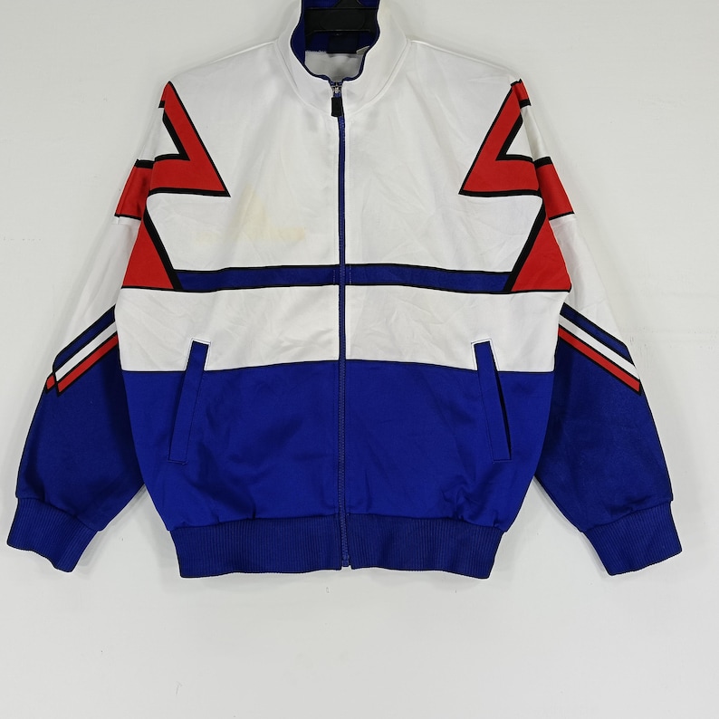May include: A white, blue, and red track jacket with a zipper closure. The jacket has a geometric pattern with a white background and blue and red accents.