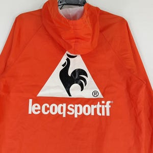 K&ouml;nnte beinhalten: Leuchtend orangefarbene Kapuzenjacke mit dem Le Coq Sportif Logo auf dem R&uuml;cken. Das Logo zeigt einen schwarzen Hahn in einem wei&szlig;en Dreieck, darunter der Markenname. Die Jacke hat eine Kapuze und ein wei&szlig;es Innenfutter.