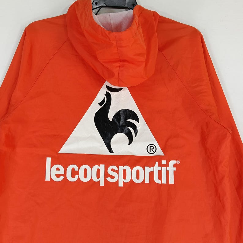 K&ouml;nnte beinhalten: Leuchtend orangefarbene Kapuzenjacke mit dem Le Coq Sportif Logo auf dem R&uuml;cken. Das Logo zeigt einen schwarzen Hahn in einem wei&szlig;en Dreieck, darunter der Markenname. Die Jacke hat eine Kapuze und ein wei&szlig;es Innenfutter.