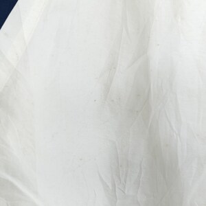 May include: Close-up of a white fabric with visible wrinkles and slight discoloration. A dark blue section is visible in the upper left corner. The fabric appears to be thin and possibly part of a garment or textile.