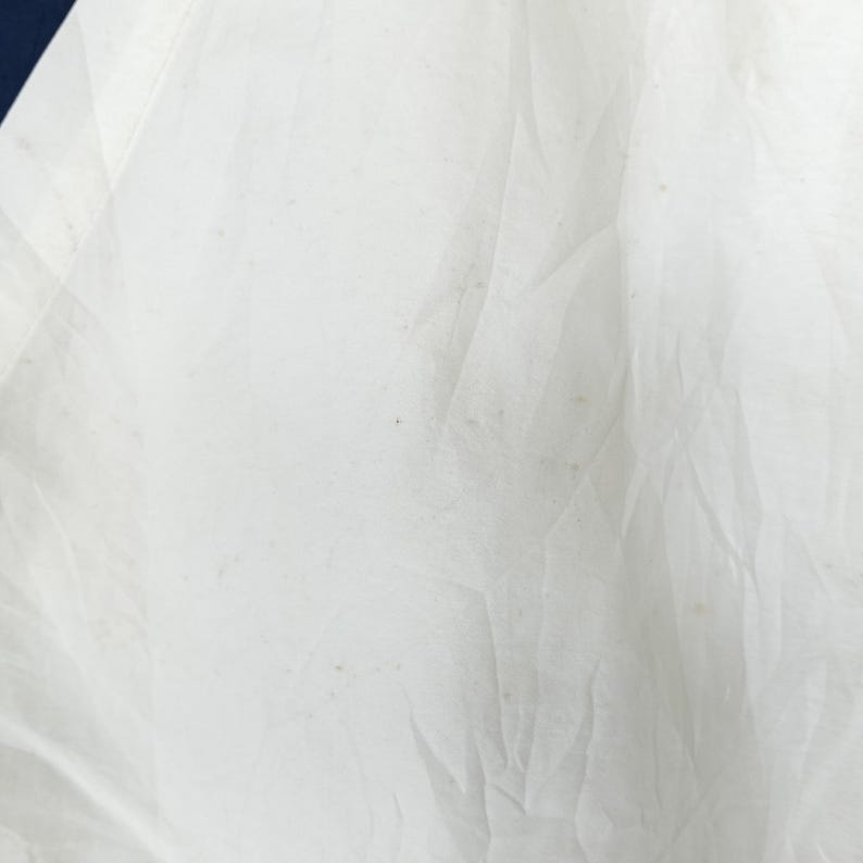 May include: Close-up of a white fabric with visible wrinkles and slight discoloration. A dark blue section is visible in the upper left corner. The fabric appears to be thin and possibly part of a garment or textile.