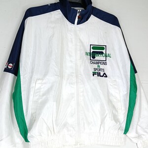 May include: White, navy blue, and green Fila windbreaker jacket. The jacket features a full zipper, a stand-up collar, and the Fila logo on the chest and sleeve. The text on the chest reads "INTERNATIONAL CHAMPIONS IN SPORTS".