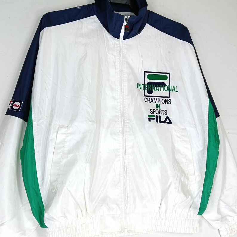 May include: White, navy blue, and green Fila windbreaker jacket. The jacket features a full zipper, a stand-up collar, and the Fila logo on the chest and sleeve. The text on the chest reads "INTERNATIONAL CHAMPIONS IN SPORTS".