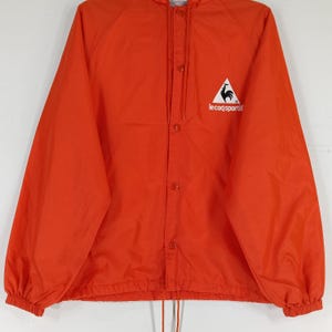 K&ouml;nnte beinhalten: Leuchtend orangefarbene Le Coq Sportif Jacke mit Kapuze und Druckknopfverschluss. Mit einem wei&szlig;en Dreieckslogo mit einem Hahn-Emblem und dem Markennamen. Die Jacke hat einen Kordelzug am Saum und elastische B&uuml;ndchen.