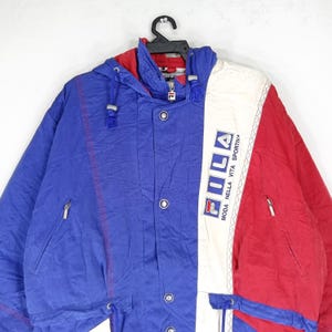 May include: A vintage Fila jacket in blue, red, and white. The jacket features a hood, zipper, and button closures. The Fila logo is prominently displayed on the white panel. The jacket has a retro, sporty aesthetic.