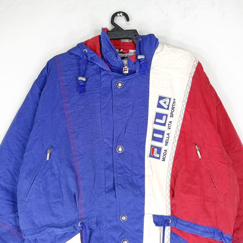 May include: A vintage Fila jacket in blue, red, and white. The jacket features a hood, zipper, and button closures. The Fila logo is prominently displayed on the white panel. The jacket has a retro, sporty aesthetic.