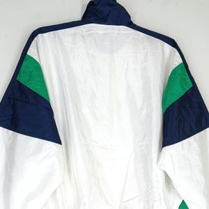 May include: A white, navy blue, and green athletic jacket. The jacket has a high collar and a full zipper. The sleeves and shoulders feature color-blocked panels of navy blue and green. The jacket is made of a lightweight, possibly nylon, material.