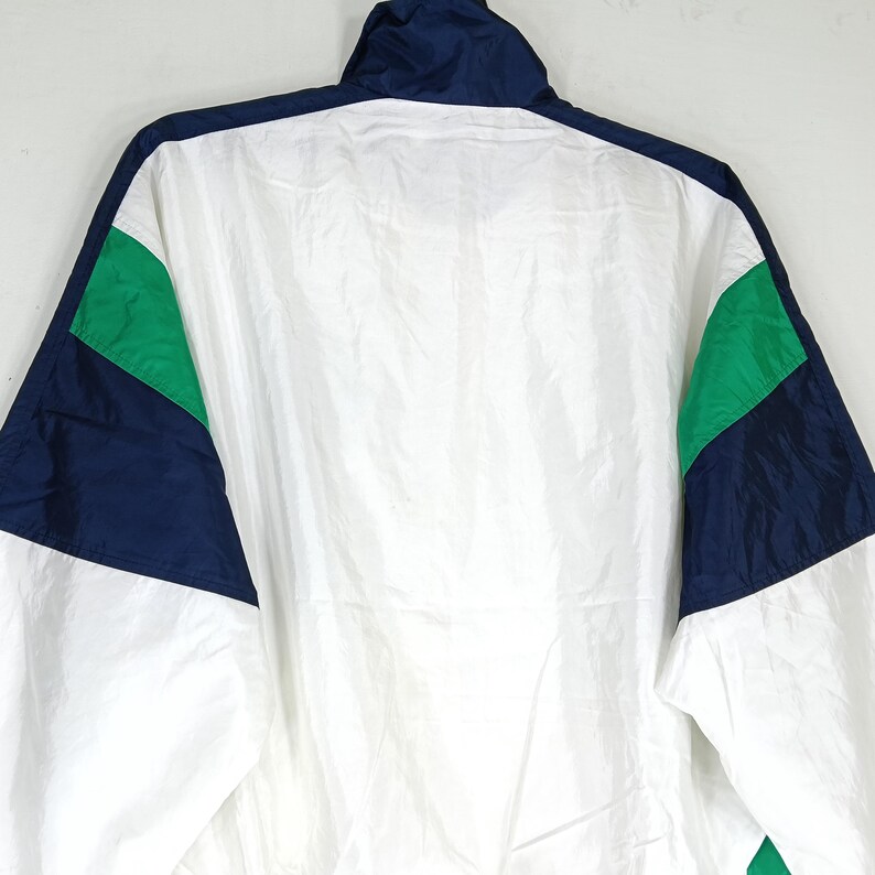 May include: A white, navy blue, and green athletic jacket. The jacket has a high collar and a full zipper. The sleeves and shoulders feature color-blocked panels of navy blue and green. The jacket is made of a lightweight, possibly nylon, material.