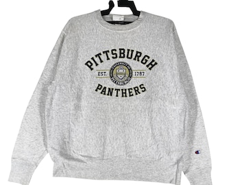 May include: A light gray sweatshirt with the text "PITTSBURGH" arched across the top, "PANTHERS" below, and a circular emblem in the center. The sweatshirt has long sleeves and a small Champion logo on the left sleeve.