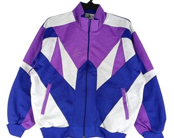 May include: A vintage track jacket with a colour block design. The jacket features a zip closure, a stand-up collar, and a geometric pattern in purple, white, and blue. The jacket has a logo on the chest and ribbed cuffs and hem.