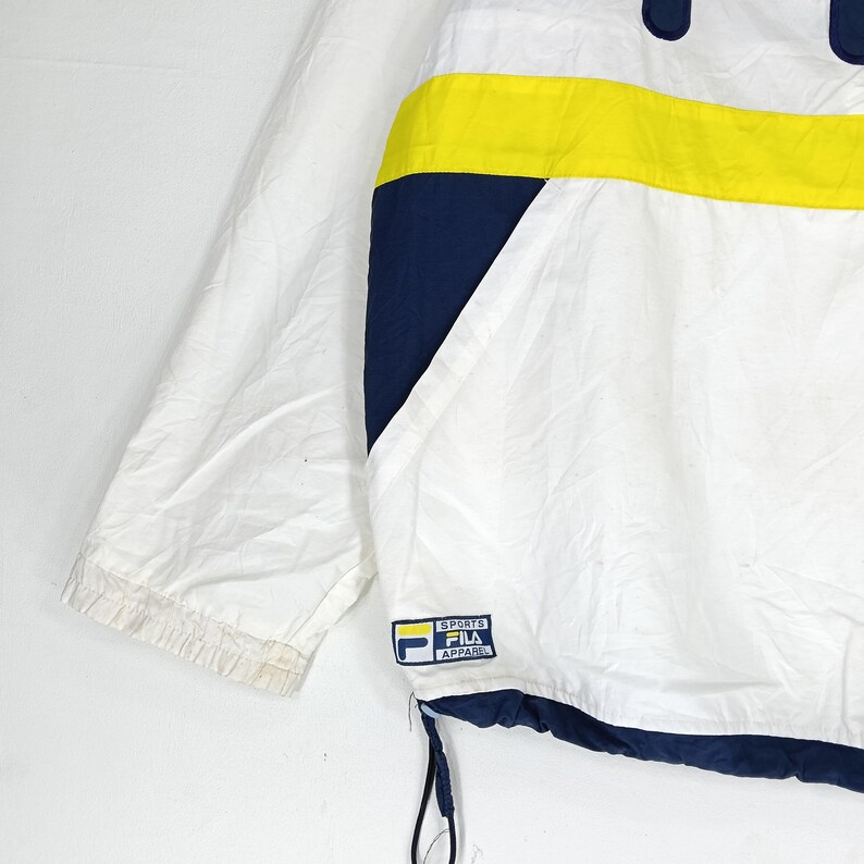 May include: White and navy blue FILA sports apparel jacket with a yellow stripe across the chest. The jacket has a drawstring at the bottom and a small FILA logo patch.