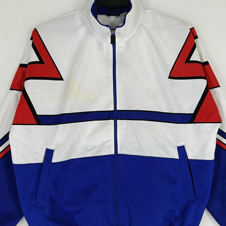 May include: A white and blue track jacket with red and black geometric designs. The jacket has a zipper closure and two pockets.