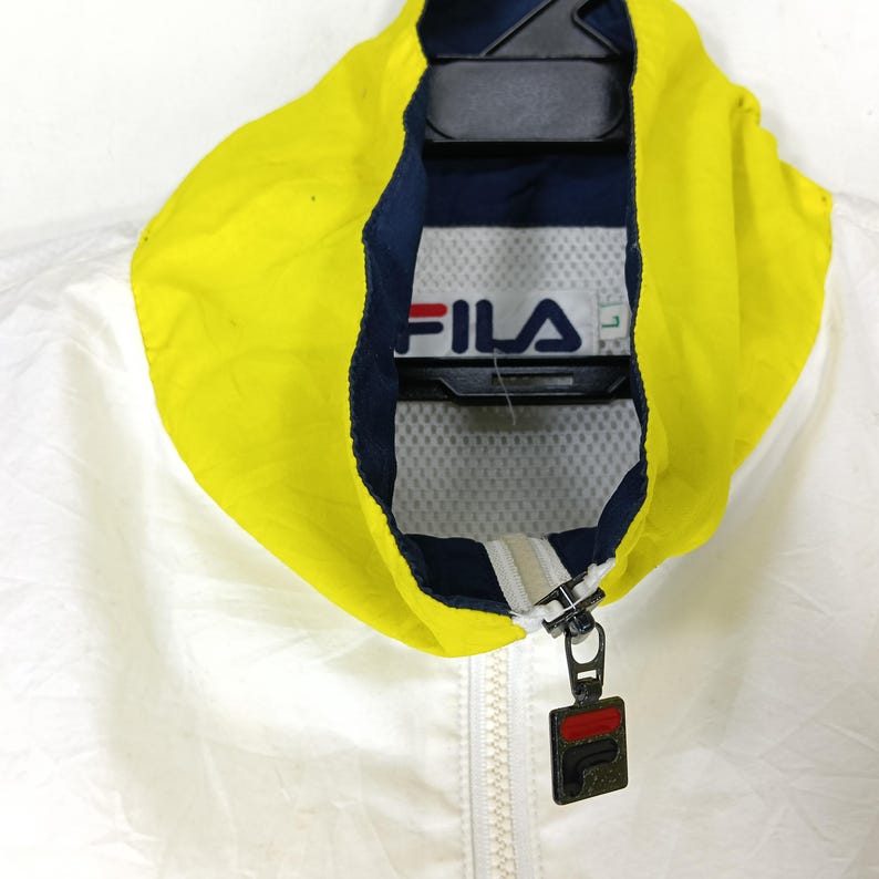 May include: A white and yellow Fila jacket with a navy blue interior. The jacket has a full zipper and a hood. The Fila logo is visible on the inside of the collar. The jacket is hanging on a black hanger.