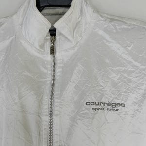 May include: A white, quilted jacket with a stand-up collar and a full-length silver zipper. The jacket features the text "courr&egrave;ges sport futur" in gray lettering on the right chest. The jacket is hanging on a black hanger.