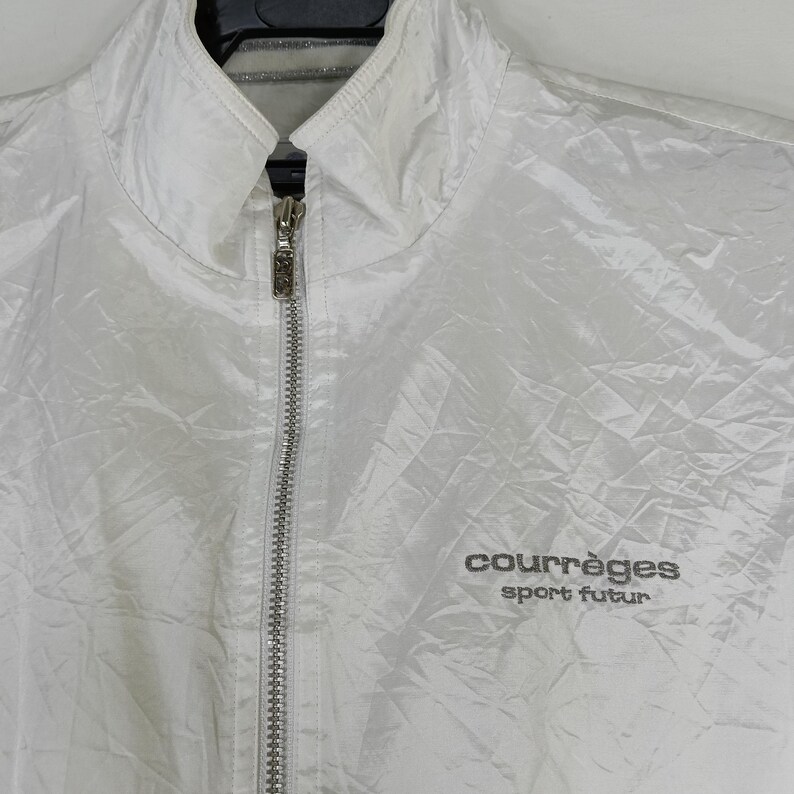 May include: A white, quilted jacket with a stand-up collar and a full-length silver zipper. The jacket features the text "courr&egrave;ges sport futur" in gray lettering on the right chest. The jacket is hanging on a black hanger.