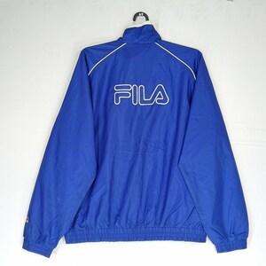 May include: A blue Fila track jacket with a white logo on the back. The jacket has a stand-up collar, white trim, and a drawstring hem. The Fila logo is in white, and the jacket is made of a lightweight material.