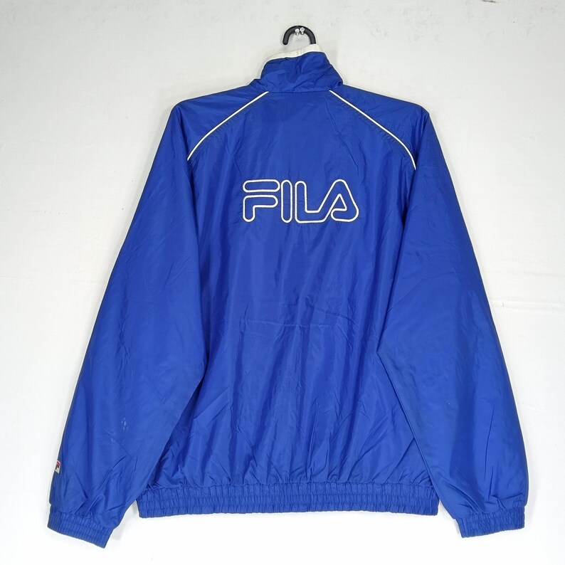May include: A blue Fila track jacket with a white logo on the back. The jacket has a stand-up collar, white trim, and a drawstring hem. The Fila logo is in white, and the jacket is made of a lightweight material.
