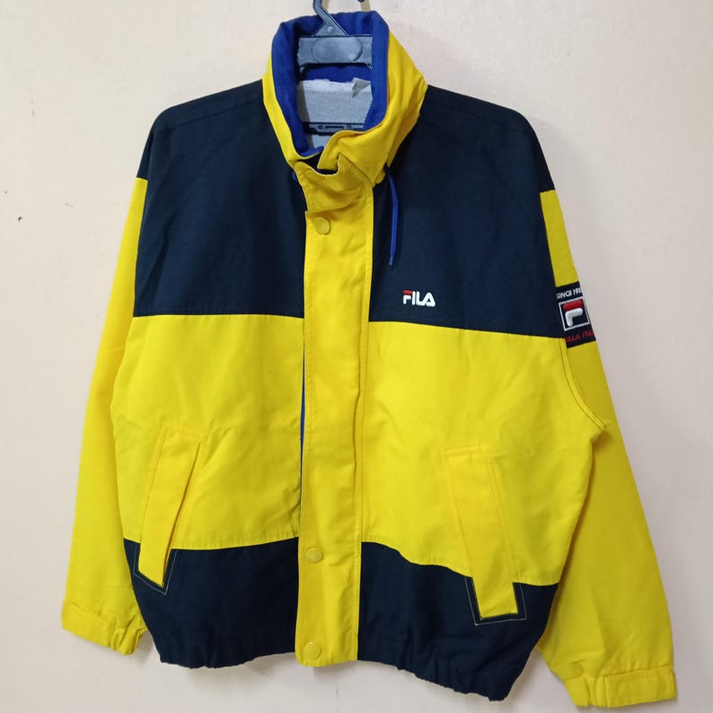 Puede incluir: Chaqueta Fila amarilla y azul marino. La chaqueta tiene un cuello alto, una cremallera frontal y un peque&ntilde;o logotipo de Fila en el pecho. La manga tiene un parche con el logotipo de Fila y las palabras "Since 1911".