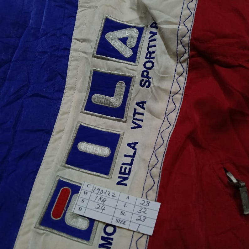 May include: Close-up of a vintage jacket featuring the FILA logo in blue and white on a cream-colored panel. The jacket has blue and red sections. The text "NELLA VITA SPORTIVA" is visible. A tag with measurements is also present.
