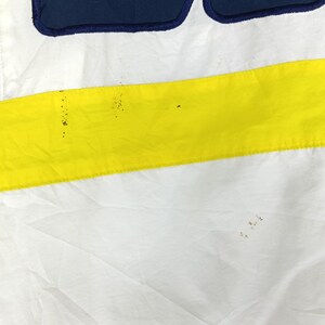 May include: Close-up of a white fabric with a horizontal yellow stripe and a navy blue section at the top. The fabric has some minor discoloration. The design is simple and bold, with a clean aesthetic.