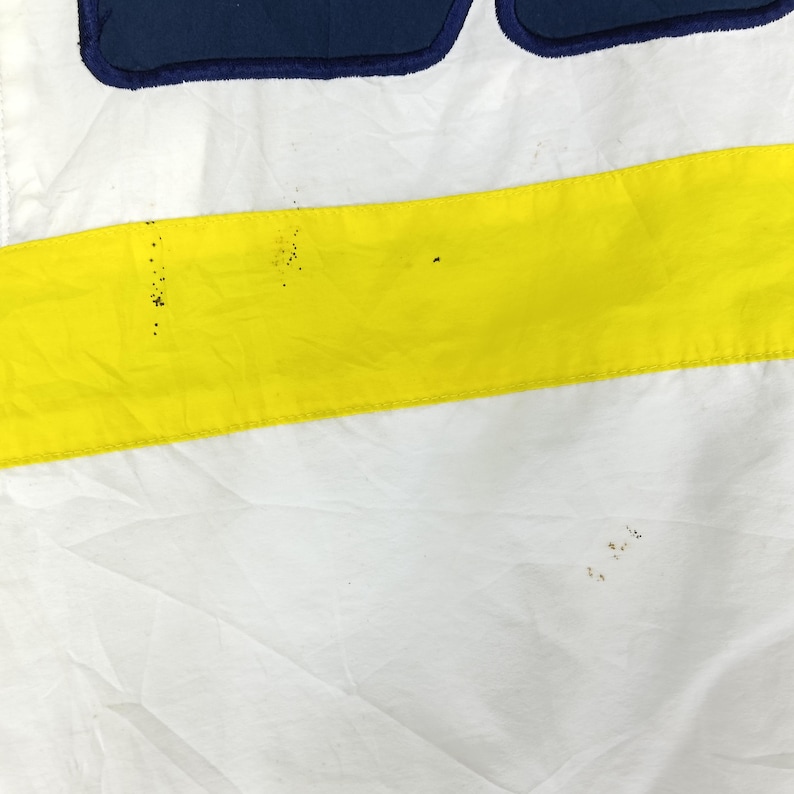May include: Close-up of a white fabric with a horizontal yellow stripe and a navy blue section at the top. The fabric has some minor discoloration. The design is simple and bold, with a clean aesthetic.